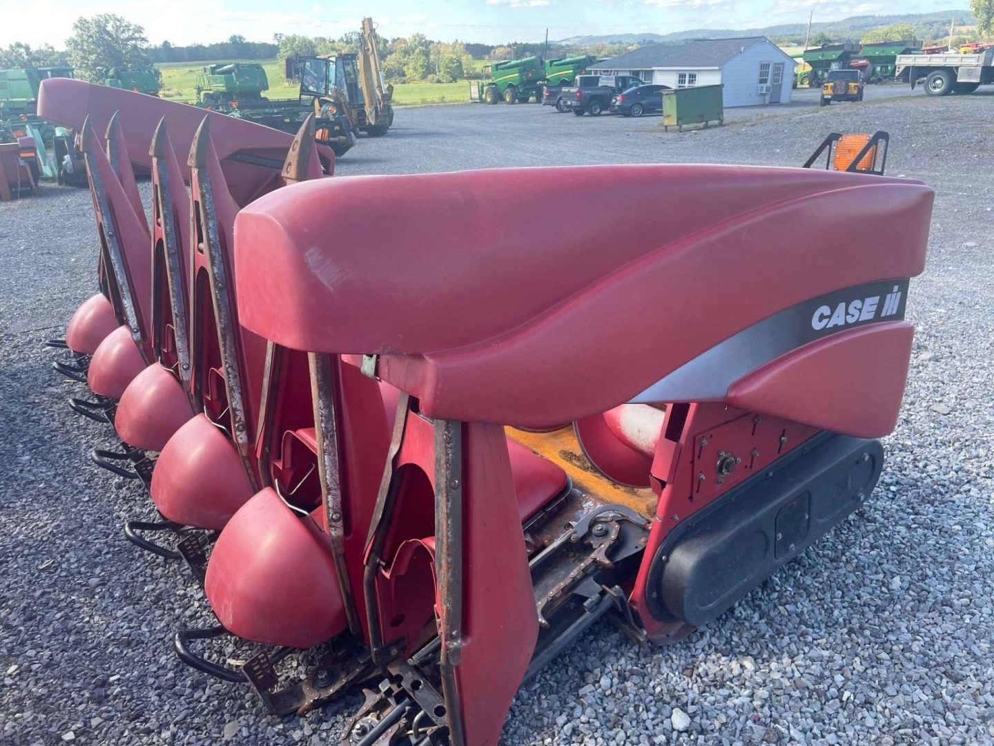 Row of red agricultural planters parked on gravel in a farmyard, with buildings in the background