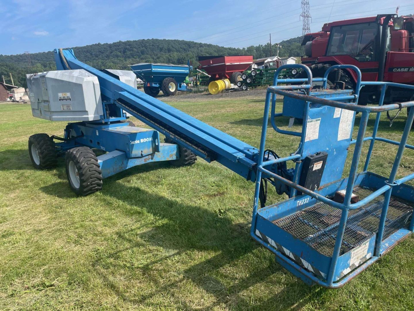 Blue boom lift parked on grass, with red truck and farm equipment in the background