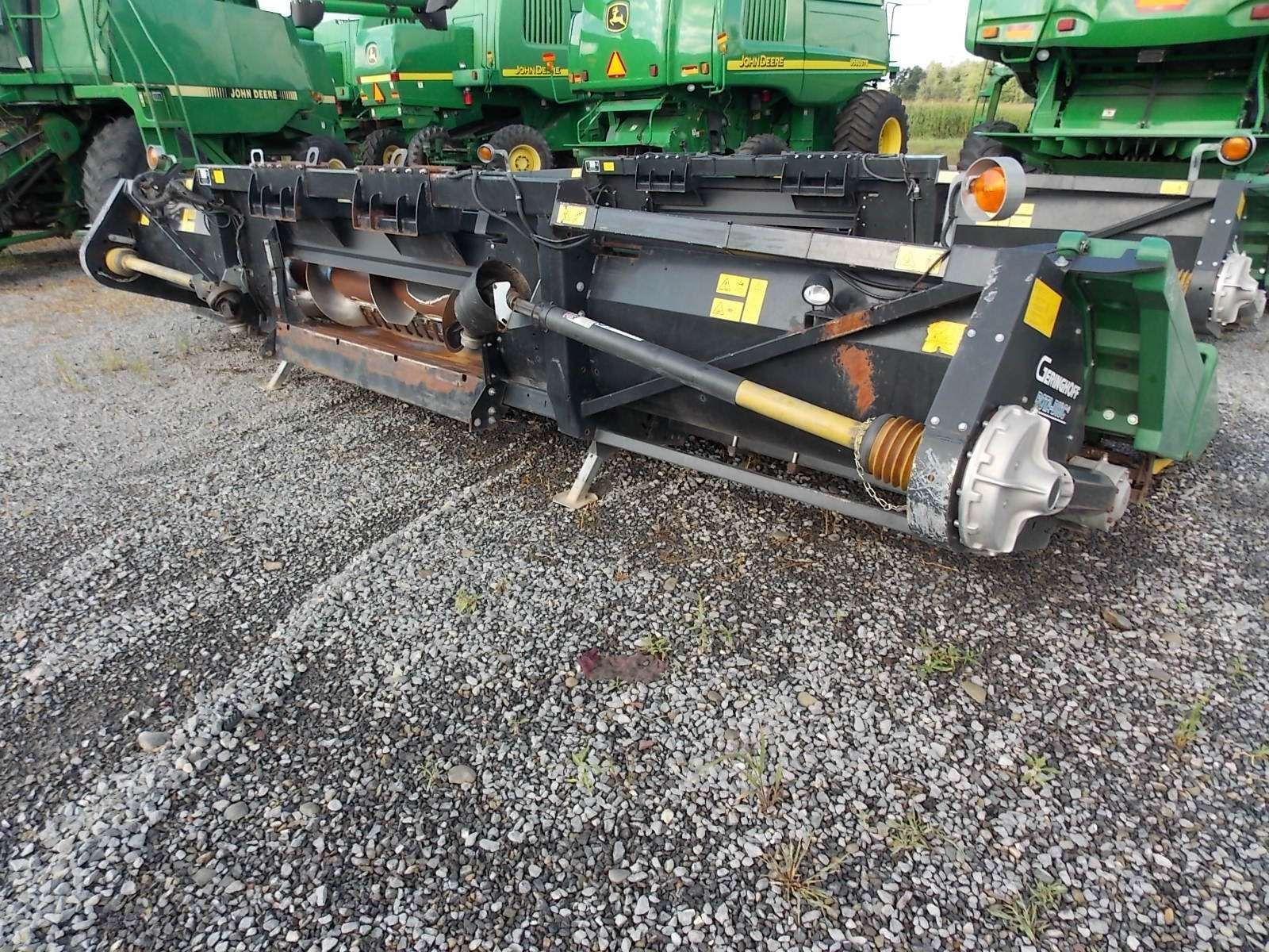 Black farm implement with orange augers parked on gravel in front of green agricultural machinery
