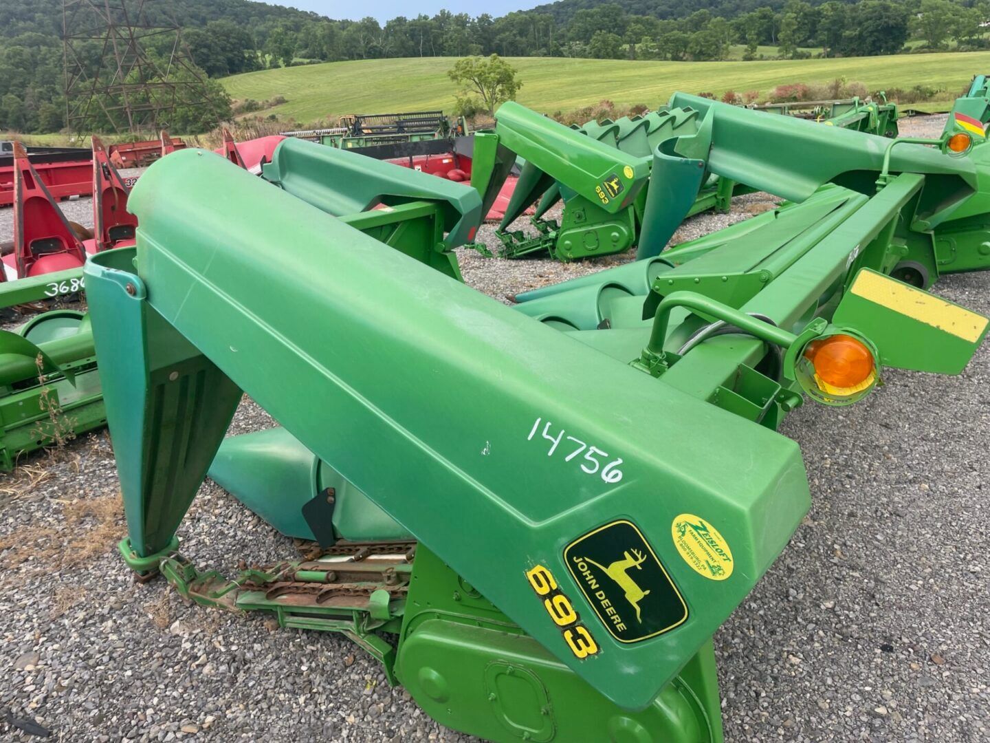 Green John Deere farm implement parked outdoors on gravel, with other machinery in the background