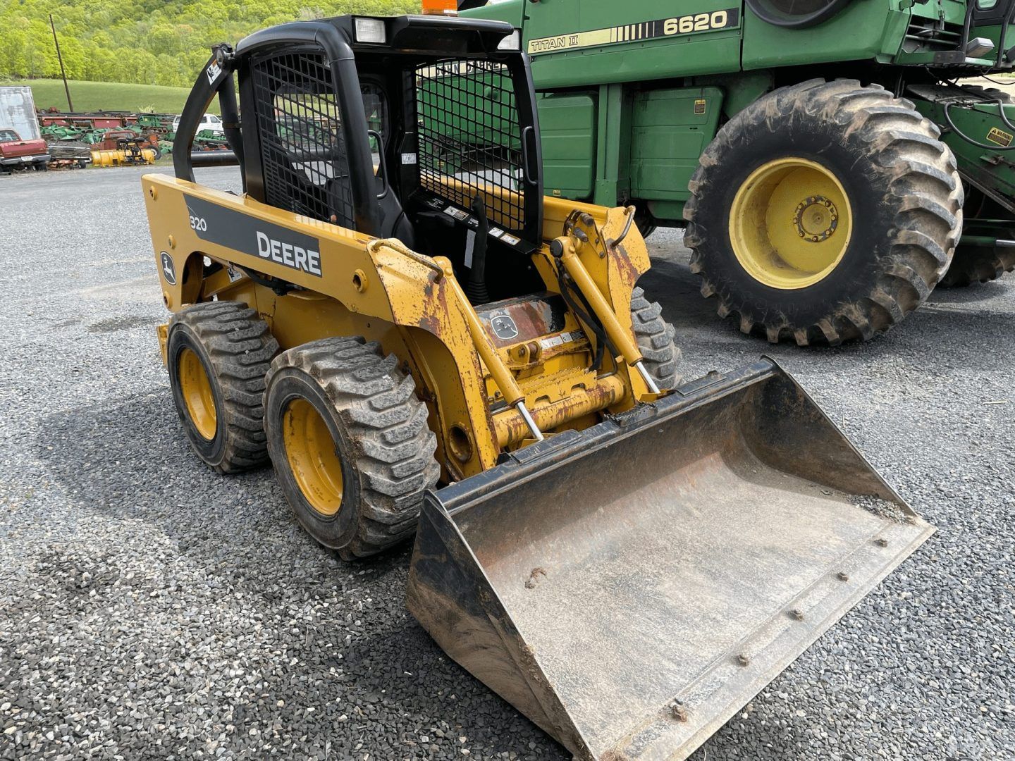 Yellow skid steer with front bucket parked on gravel beside a large green farm truck