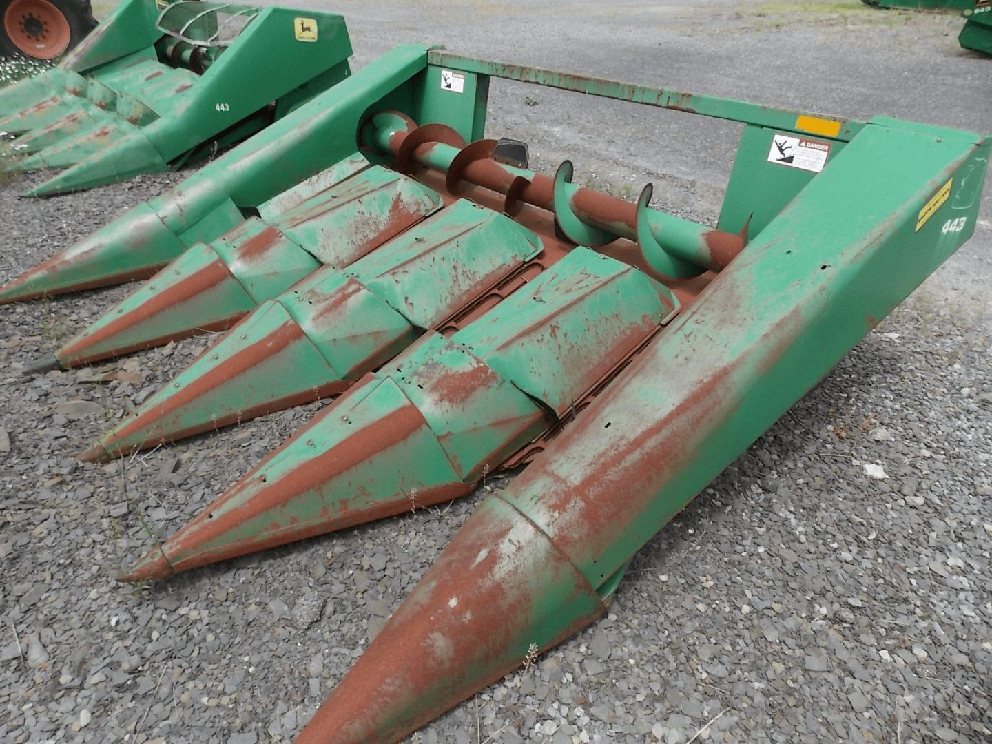 Green farm corn header attachment with rusted metal tines on gravel.