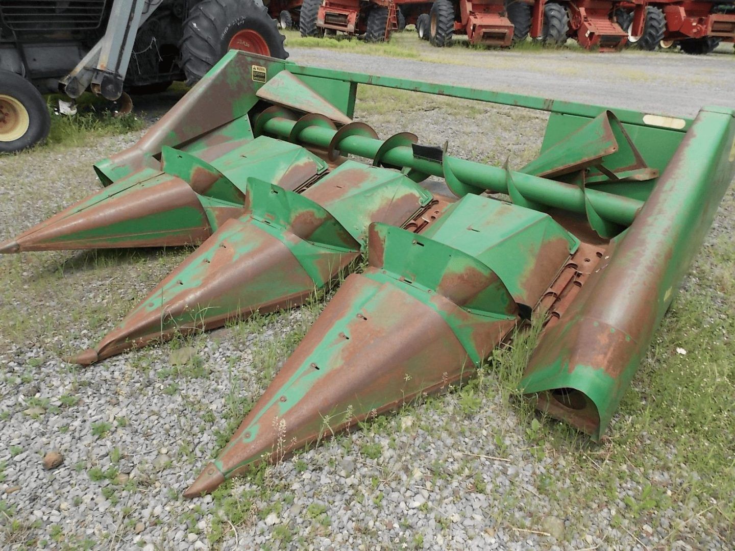 Green farm corn header attachment with pointed metal snouts, resting on gravel outdoors