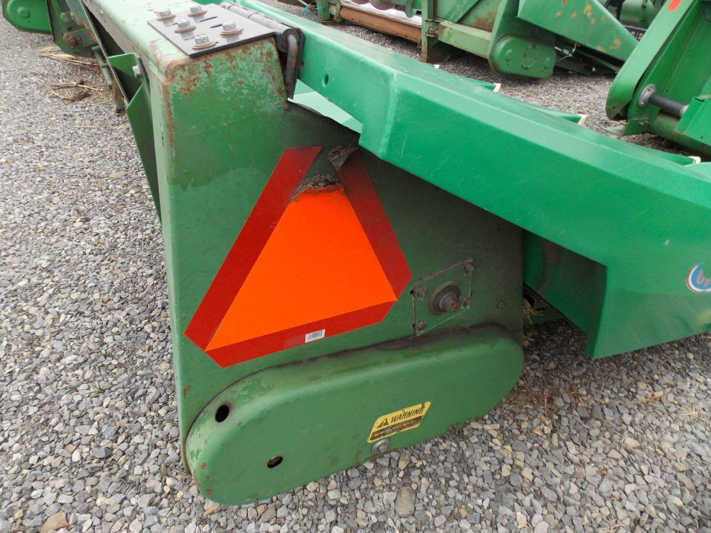 Close-up of a green farm machine with an orange reflective safety triangle on a gravel surface