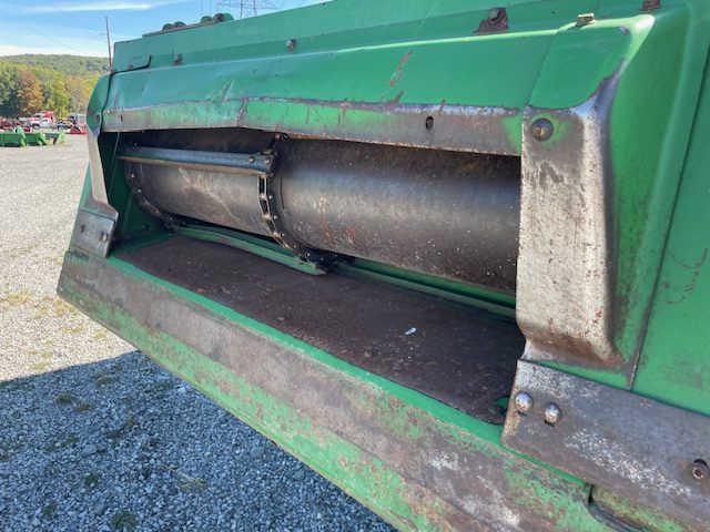 Close-up of green farm machinery with a metal drum inside, sitting outdoors on gravel.