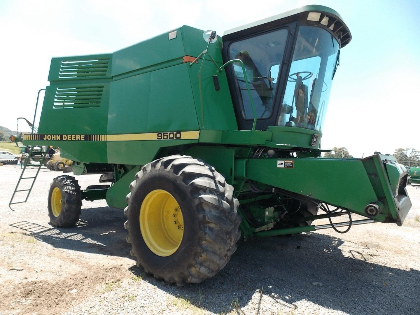 Green combine harvester with large tires parked on a gravel lot under a bright sky