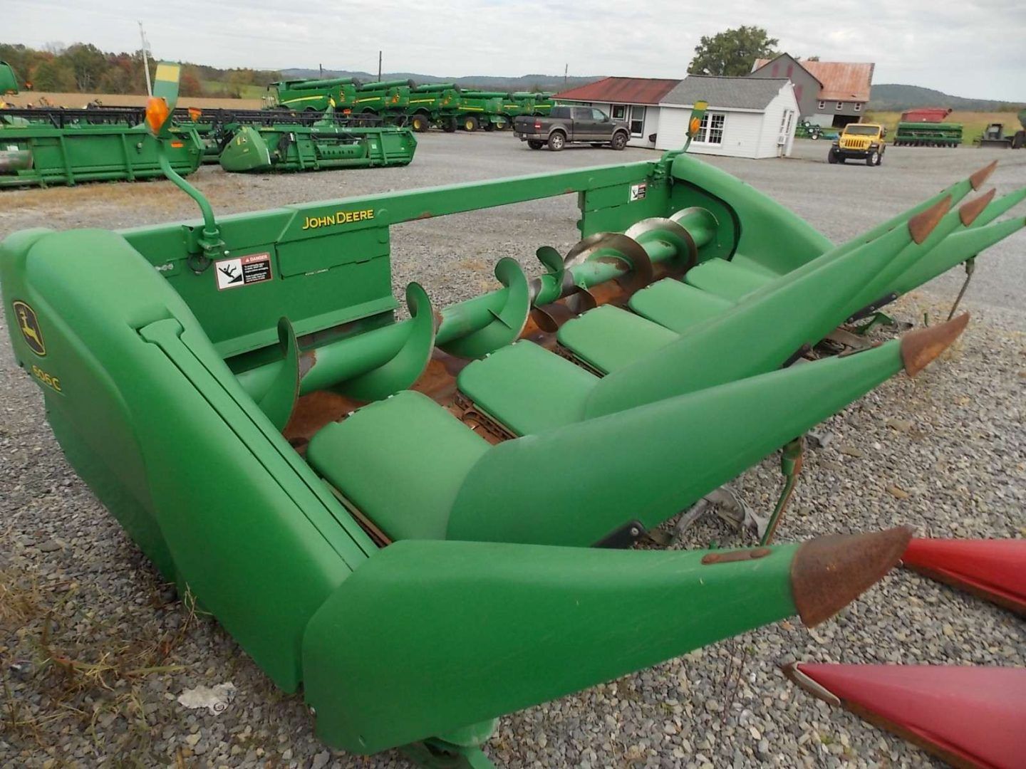 Green agricultural corn header attachment parked in a gravel lot with other farm machinery nearby