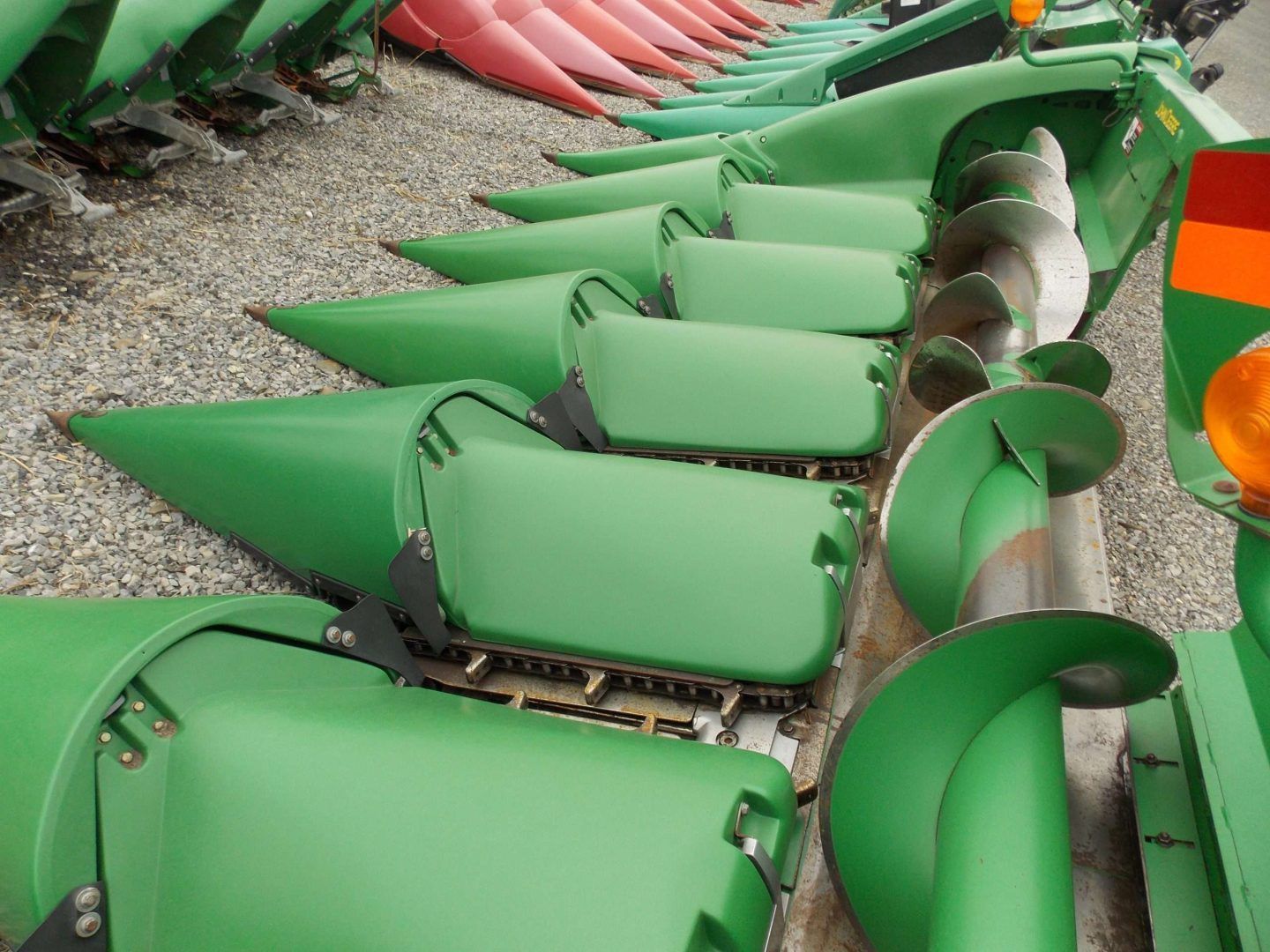 Row of green agricultural machine attachments lined up on gravel, with pink ones in the background