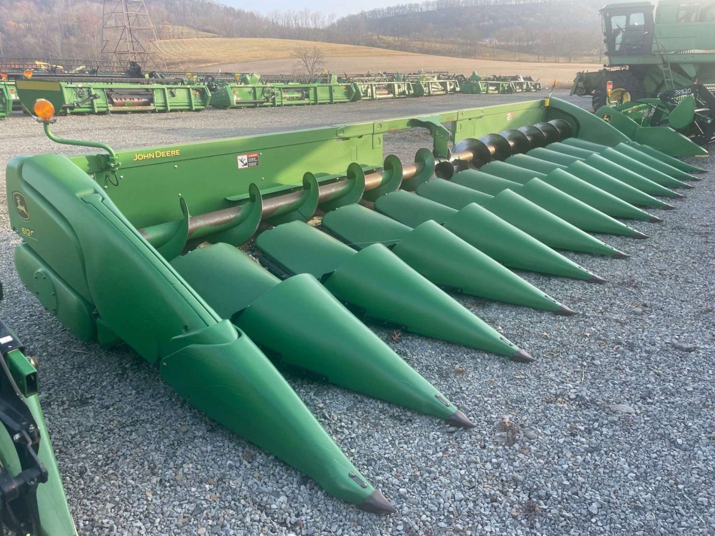Green corn header on gravel, with pointed row dividers and farm equipment in the background