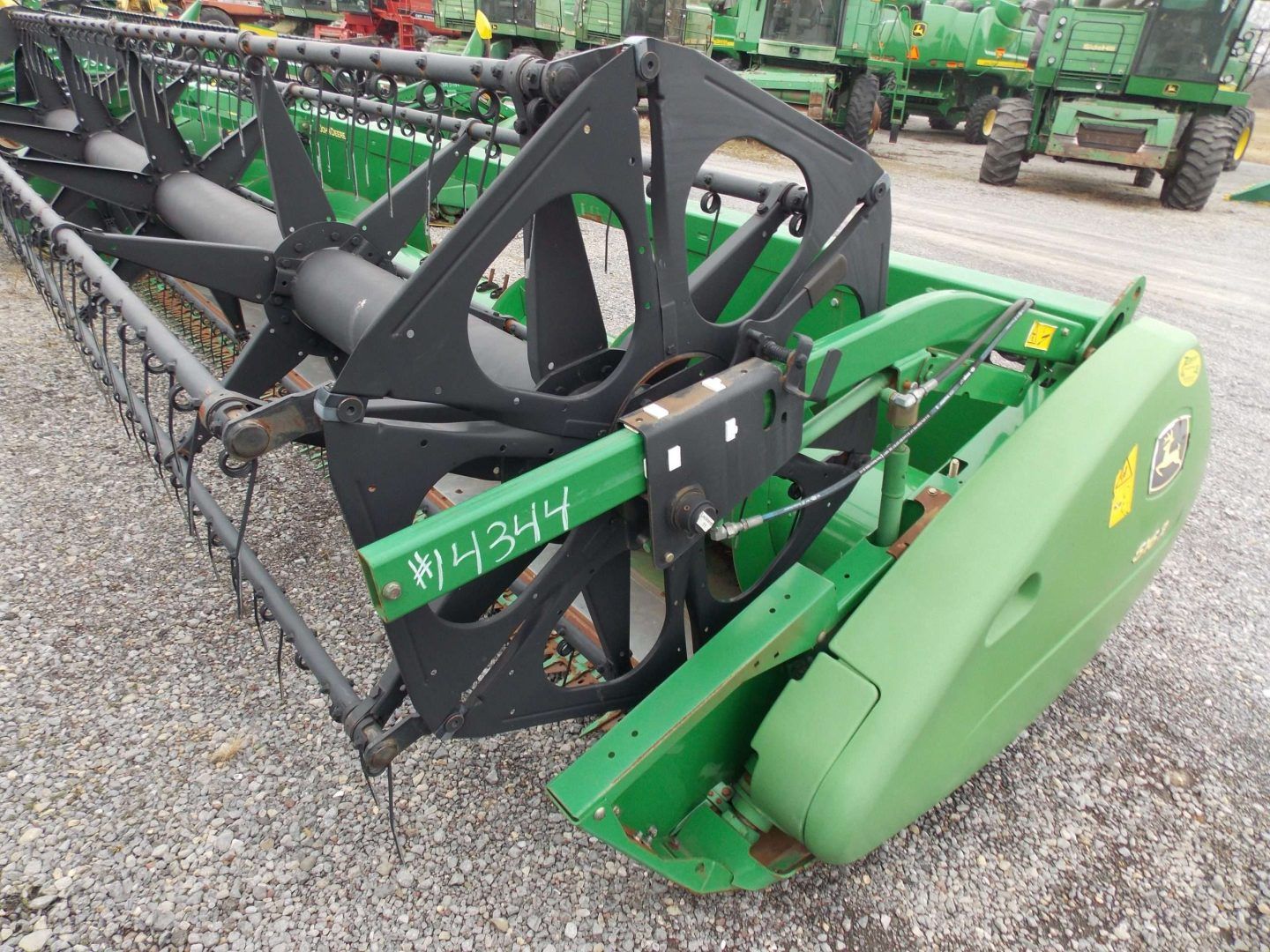 Green farm equipment with a black header attachment parked on gravel outdoors