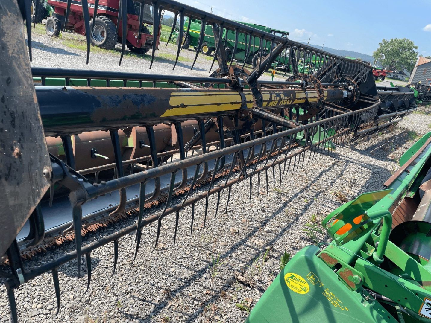 Row of farm combines with long black headers lined up on gravel lot, green tractor at right.