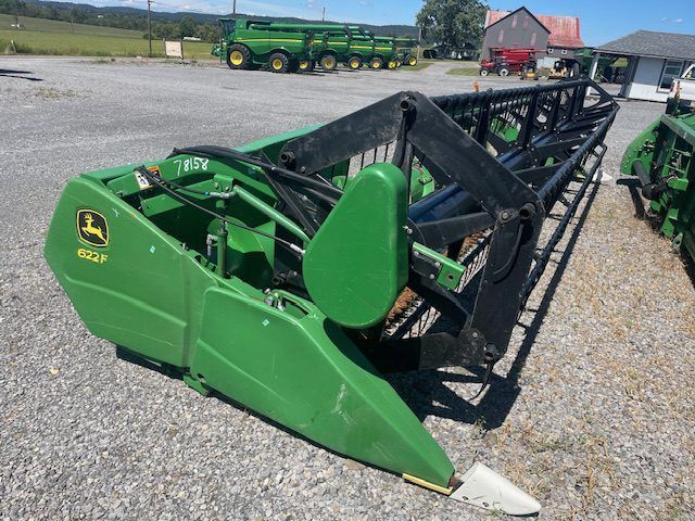 Green farm loader attachment on gravel lot, with black metal frame and parked machinery in background