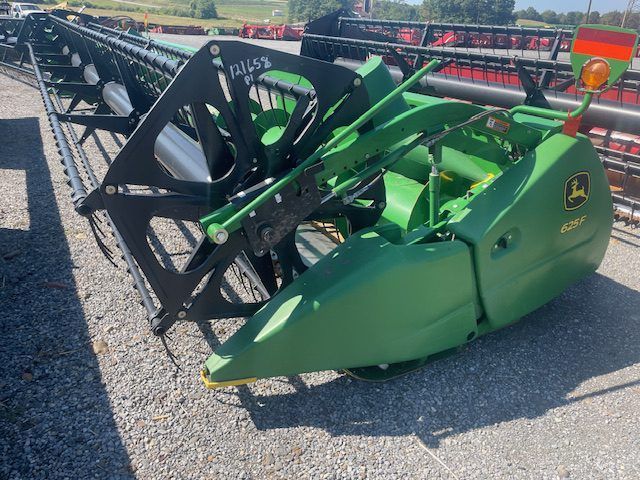 Green John Deere combine header with black reel parked outdoors on gravel near a field railing.
