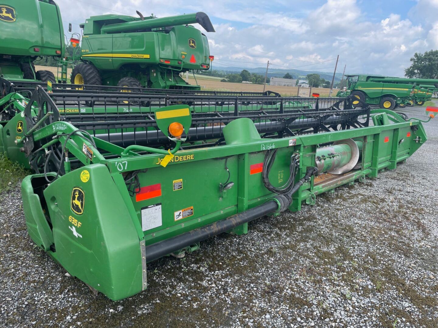 Green John Deere agricultural combine headers parked on gravel under a cloudy sky