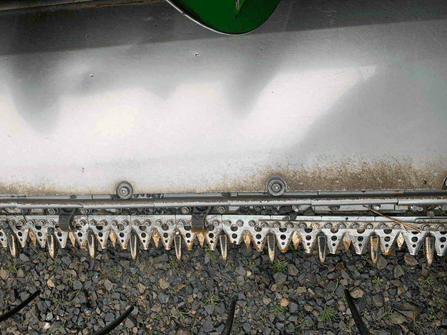 Close-up of a metal brush scraping gravel, with a green machine edge above and white dust along the blade.