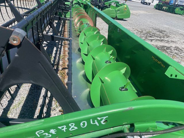 Close-up of a green farm header with a rotating auger and black guard rails in a gravel yard