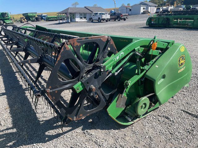 Green John Deere combine harvester header parked on gravel lot near buildings and trucks.