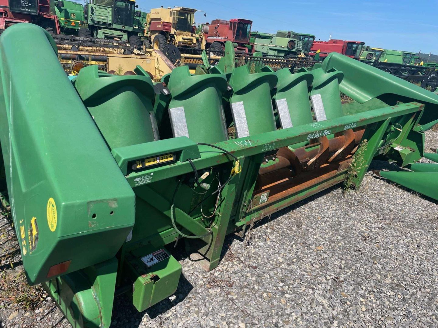 Green farm machinery with multiple cutting blades on a gravel lot, with other equipment in the background