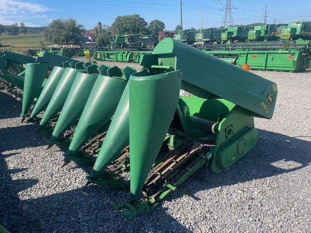 Green farm corn harvesting header with pointed metal cones, parked on gravel outdoors under blue sky