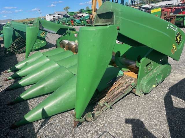 Green agricultural harvester header with pointed cones, parked outdoors on a gravel lot