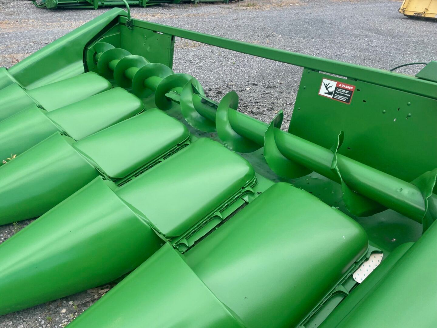 Green agricultural harvester header with metal tines and rollers on gravel.