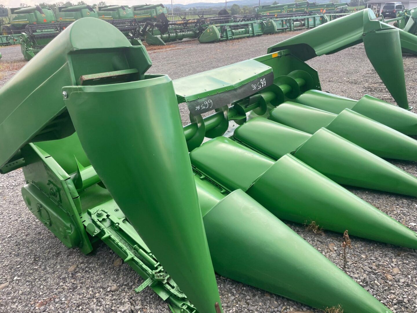 Green agricultural corn header with pointed cones, shown on gravel in an equipment yard