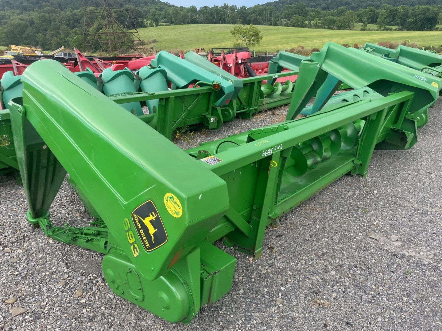 Green agricultural equipment lined up outdoors on gravel, with fields and trees in the background