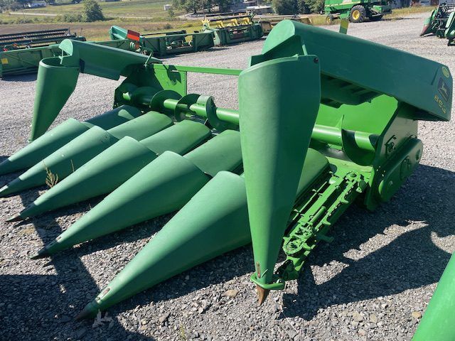 Green agricultural corn harvester with large pointed crop heads parked on gravel lot