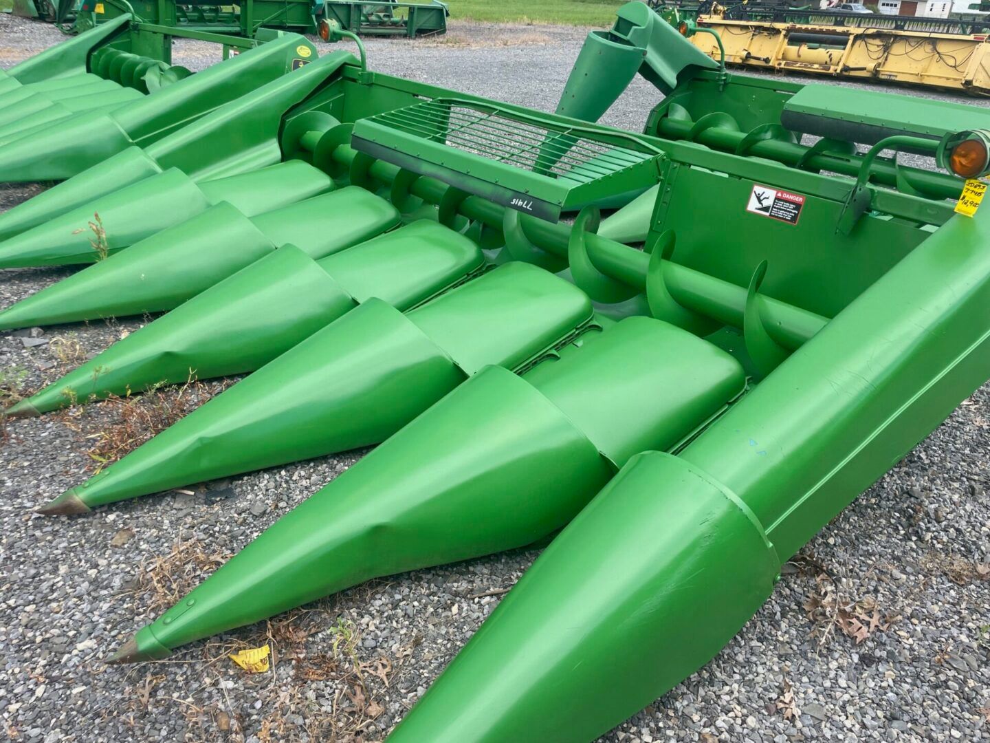 Green corn header attachment with pointed metal cones, parked on gravel outdoors