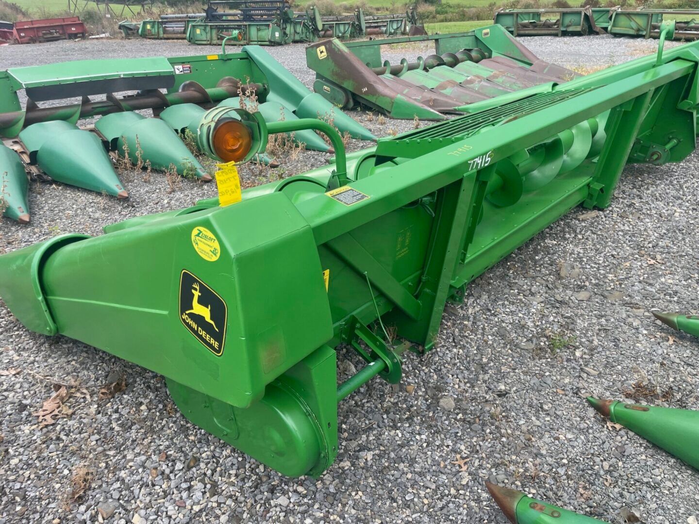 Green John Deere corn header on gravel at an equipment lot, with yellow-orange components and folded stalk snouts