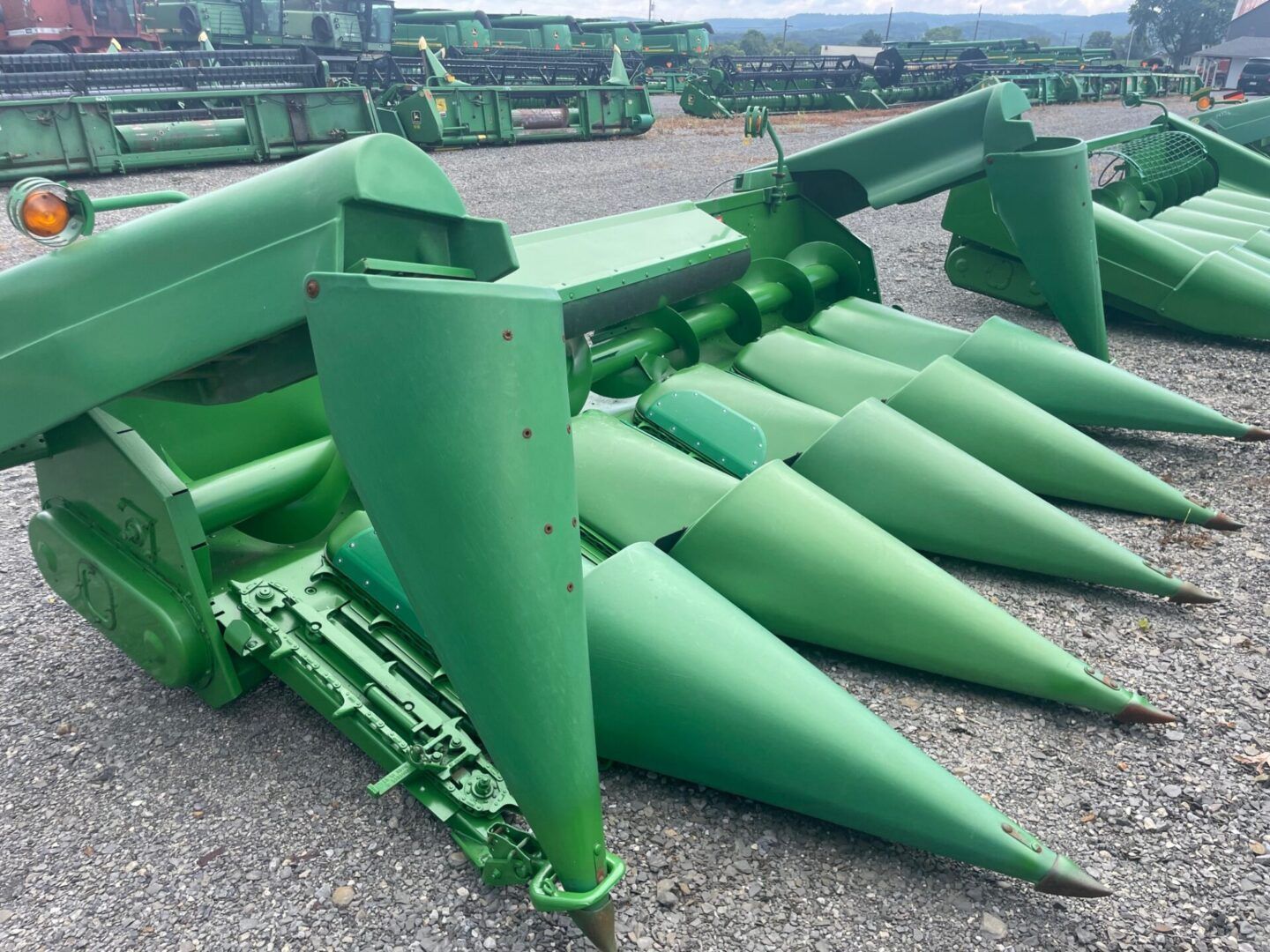 Green corn harvester header with pointed intake cones, parked on gravel at an equipment yard