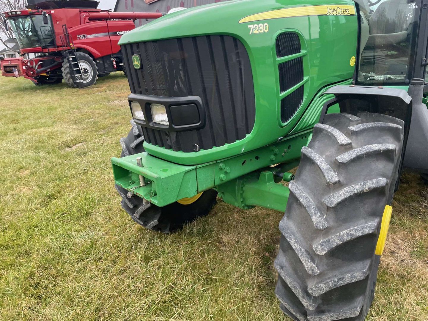 Green John Deere tractor with large front tire parked on grass, red farm equipment in the background