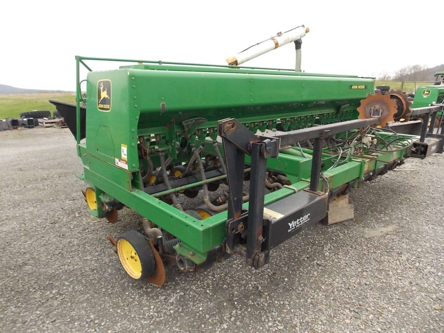 Green agricultural planter machine parked on gravel, with yellow wheels and exposed metal parts.