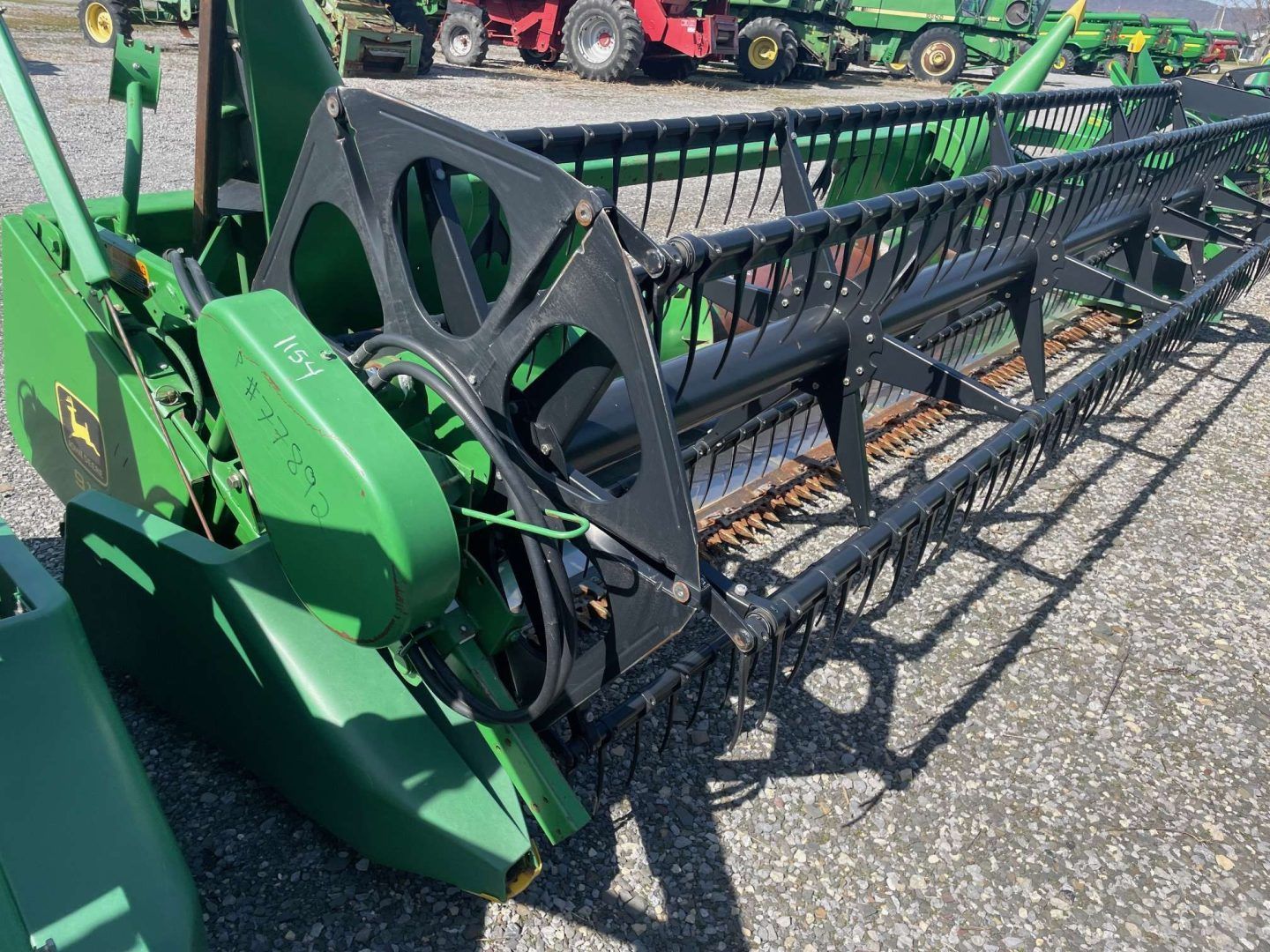 Green agricultural combine header parked on pavement, with black cutting reel and red tractor in background