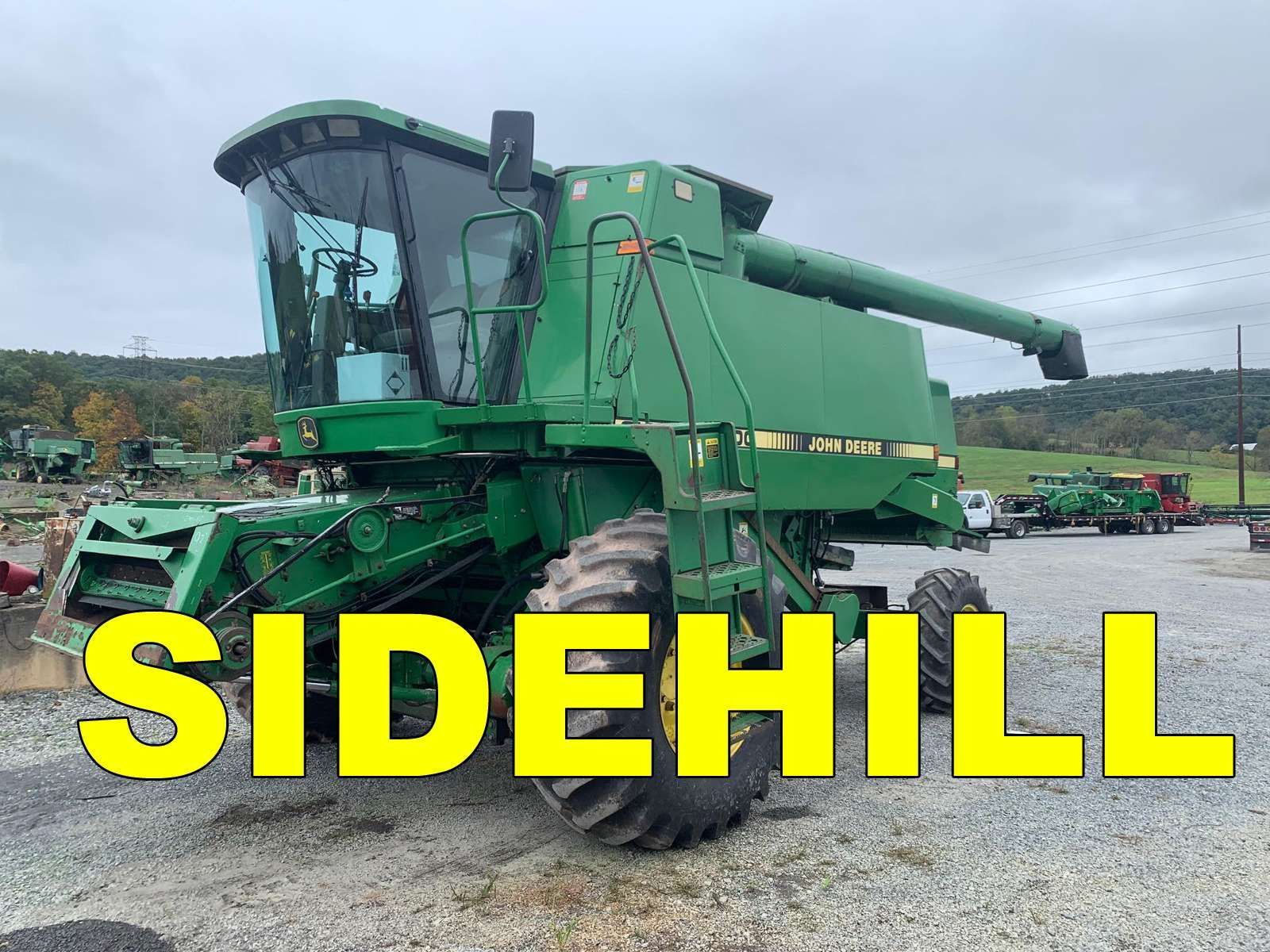 Green combine harvester labeled “SIDEHILL” parked on a gravel lot under cloudy skies