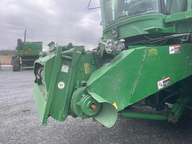 Green agricultural machine with folded attachment parked on gravel lot under cloudy sky