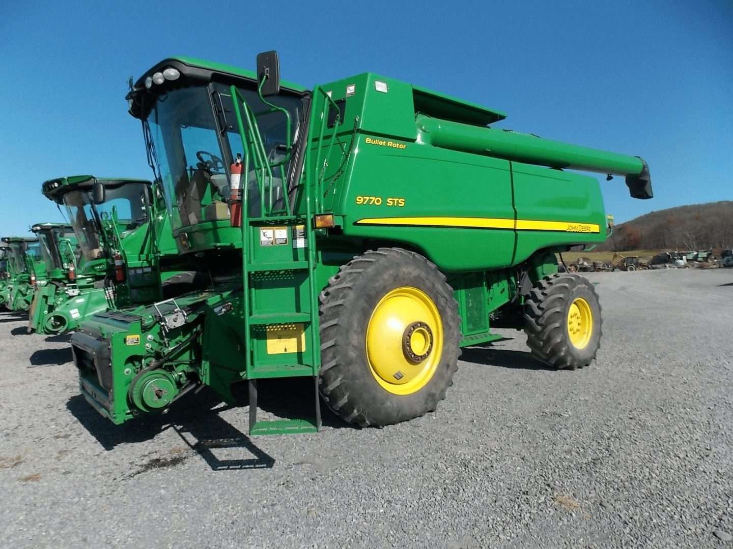 Green John Deere combine harvester on a gravel lot under a clear blue sky.