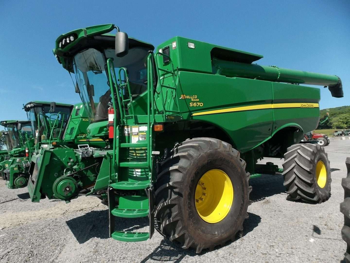 Green combine harvester with large yellow wheels parked on gravel under a blue sky.