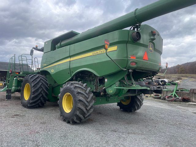 Green combine harvester with yellow wheels on a gravel lot, unloading auger extended.