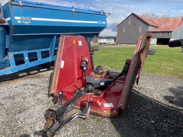 Red agricultural mower attachment on gravel beside a blue trailer and barnyard buildings