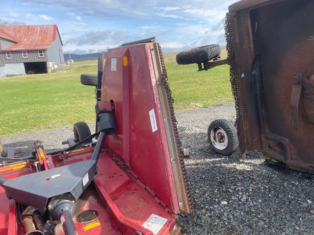 Overturned red mower deck and brown equipment on gravel near a barn and grassy field