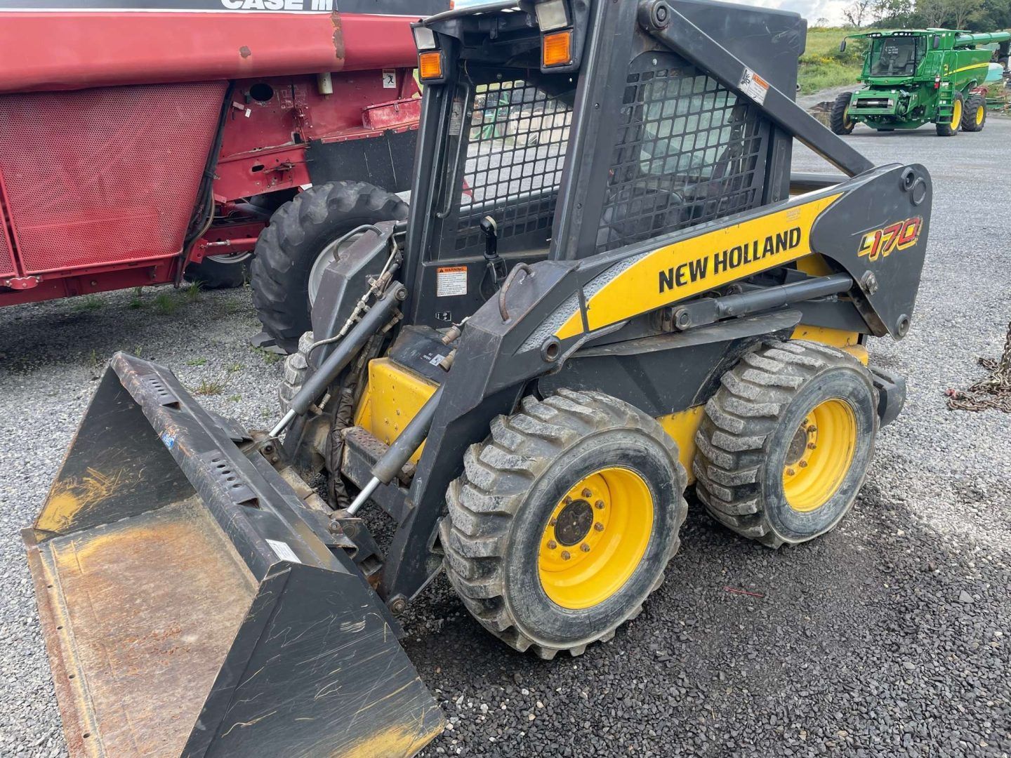 Yellow New Holland skid steer loader with front bucket parked on gravel рядом a red trailer.
