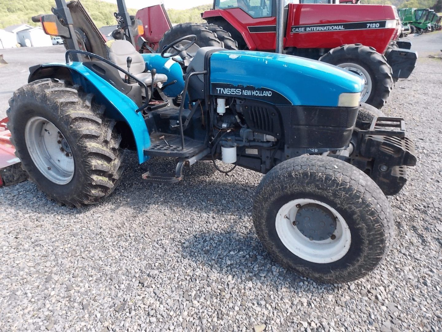 Blue tractor parked on gravel, side view, with a red truck in the background.
