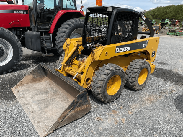 Yellow John Deere skid steer with bucket on gravel lot, beside a red tractor.