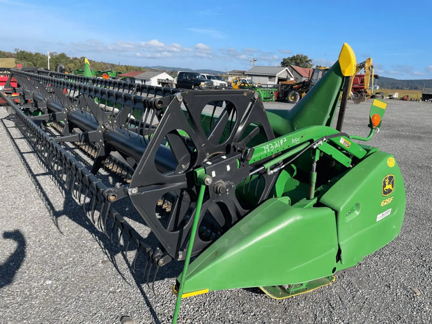 Green agricultural combine header on gravel lot under blue sky, with a worker nearby.