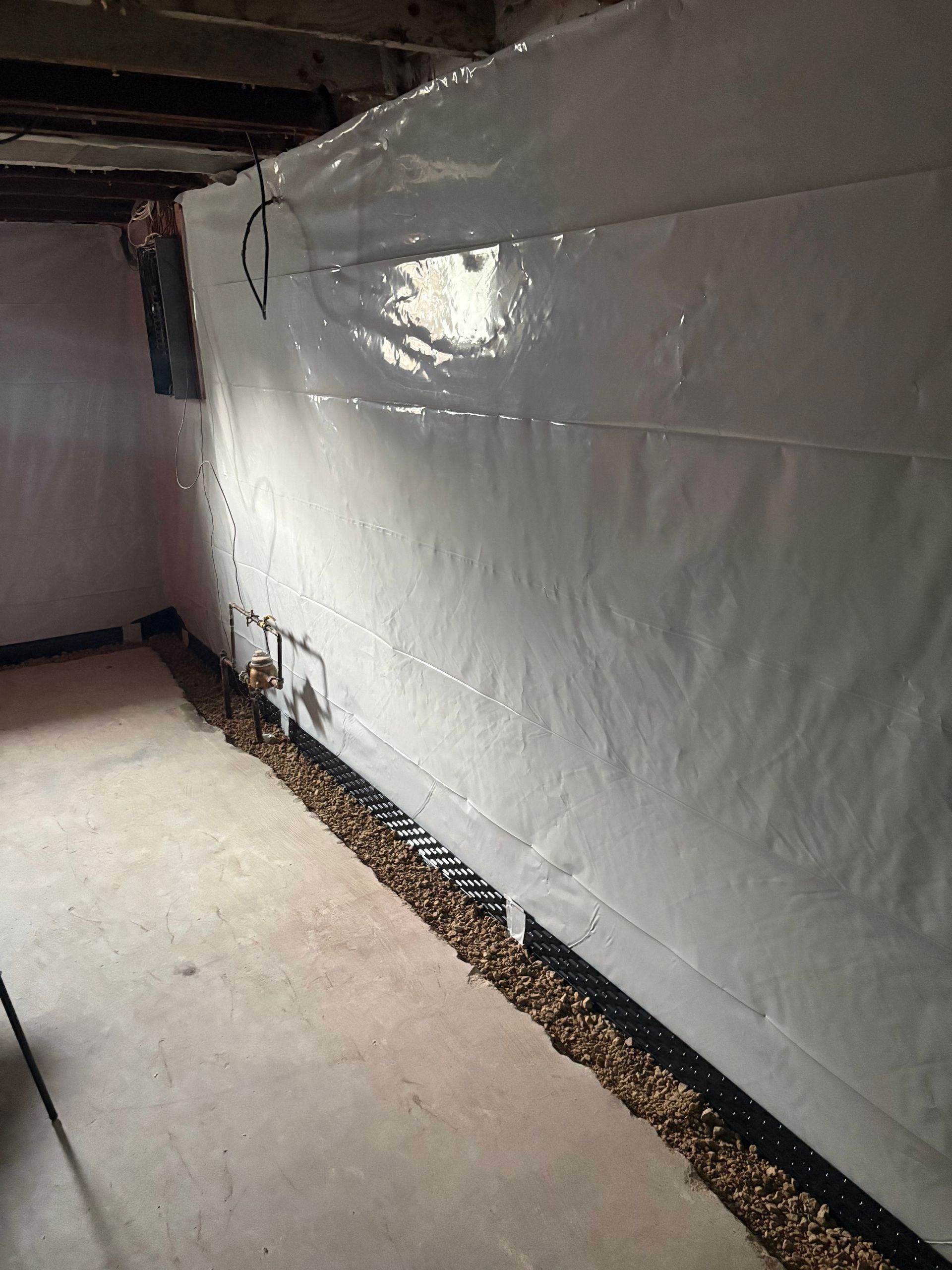 Basement wall covered with white plastic sheeting beside a concrete floor and exposed joists