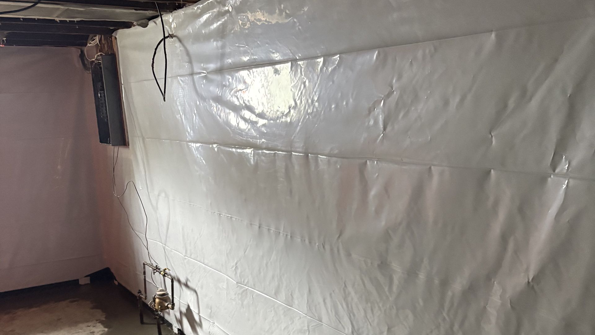 White plastic sheeting covers a basement wall, with exposed pipes and wiring near the ceiling.