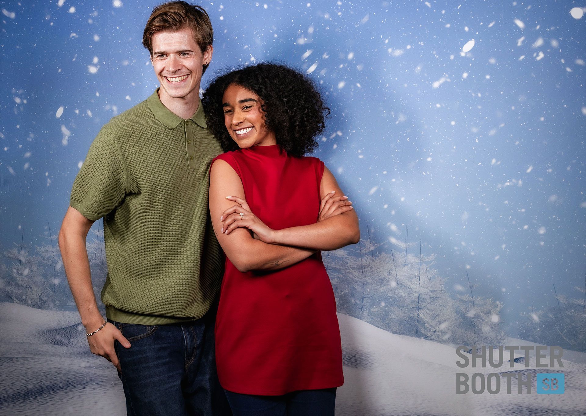 A smiling man and woman pose in front of a snowy backdrop. The woman has crossed arms and wears red. The man wears green.