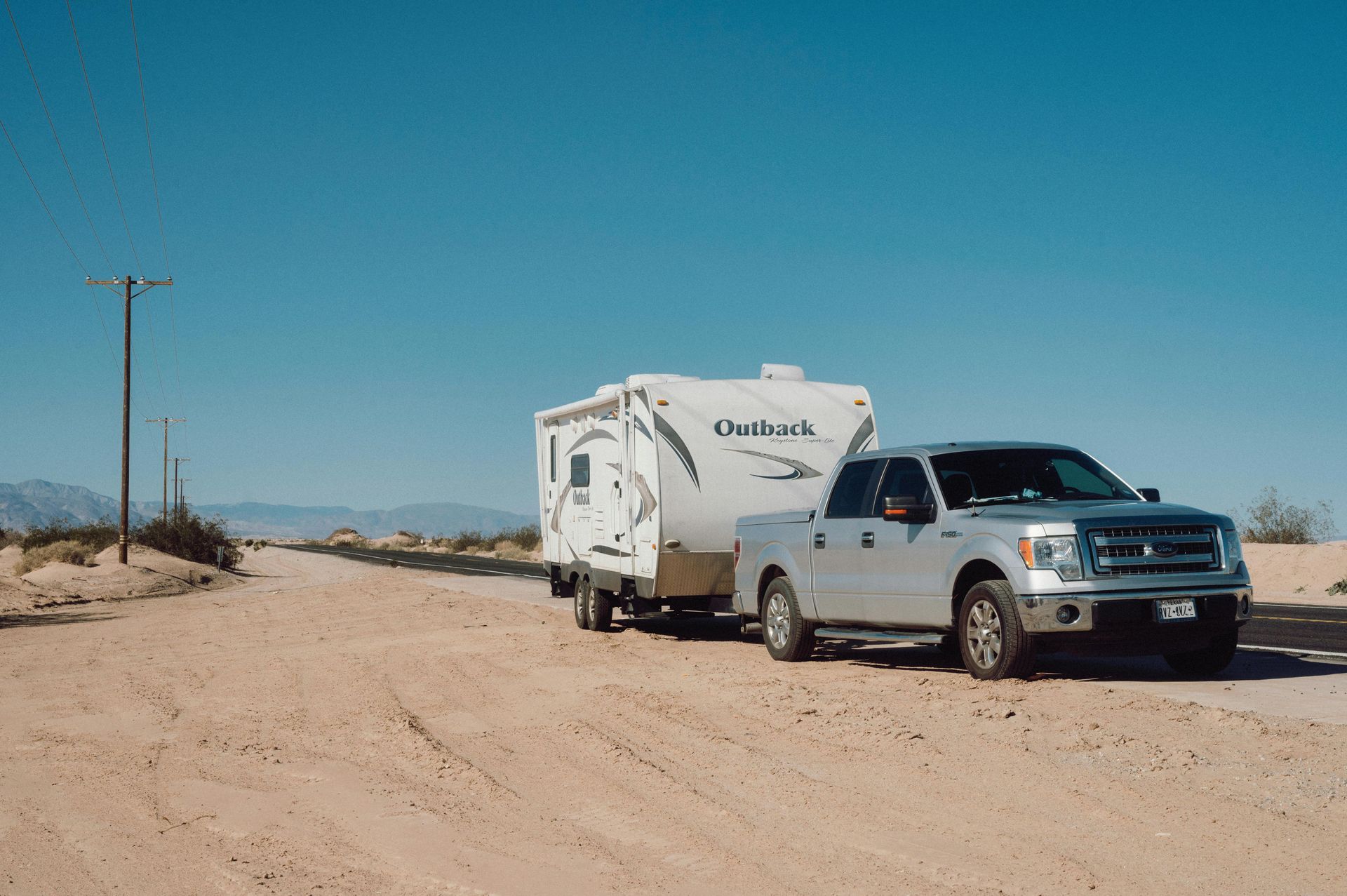 A silver truck is towing a trailer down a dirt road.