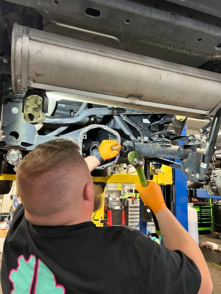 Mechanic working on a car's undercarriage, wearing orange gloves, using a tool, in a garage.